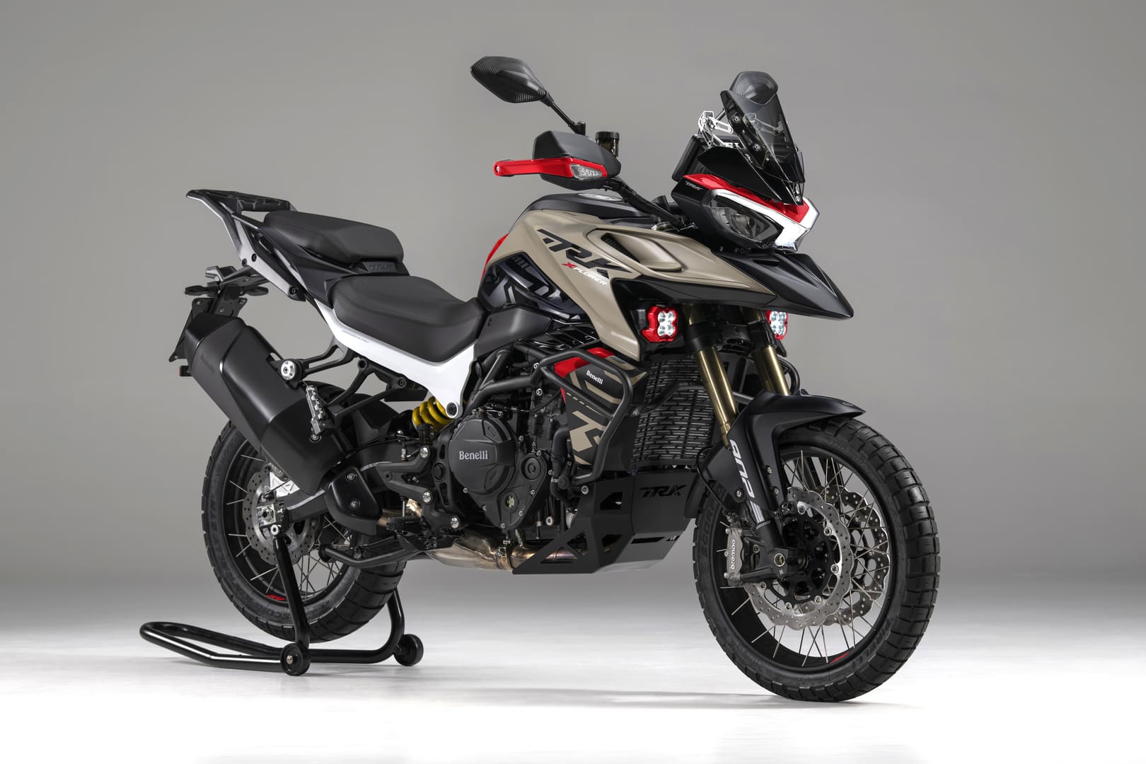 Beige ja musta Benelli TRK 902 Xplorer-moottoripyörä punaisilla yksityiskohdilla, kuvattu sivulta studioharmaata taustaa vasten.